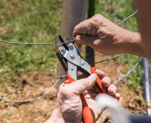 Gros plan sur des mains qui tendent et fixent un fil de clôture métallique avec une pince.